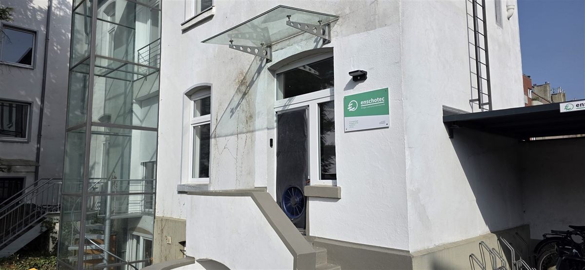 Eingang des enschotec-Büros in der Burghofstraße 40 in Düsseldorf mit grünem Firmenschild, in der Tür ein Blower-Door-Gebläse